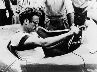 James Dean sits behind the wheel of a sports car in a still from 'The James Dean Story,' 1957. (Credit: Warner Bros./Getty Images)
