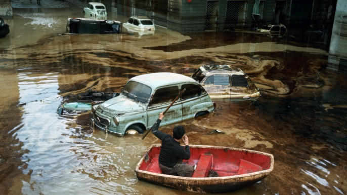 A man paddles a small boat among wrecked cars on a waterlogged street in Florence, following the devastating flood of November 1966, during which the water of the Arno rose as high as 20 feet, submerging sculpture, paintings, mosaics and manuscripts in the city's libraries. (Credit: Vittoriano Rastelli/Corbis via Getty Images)