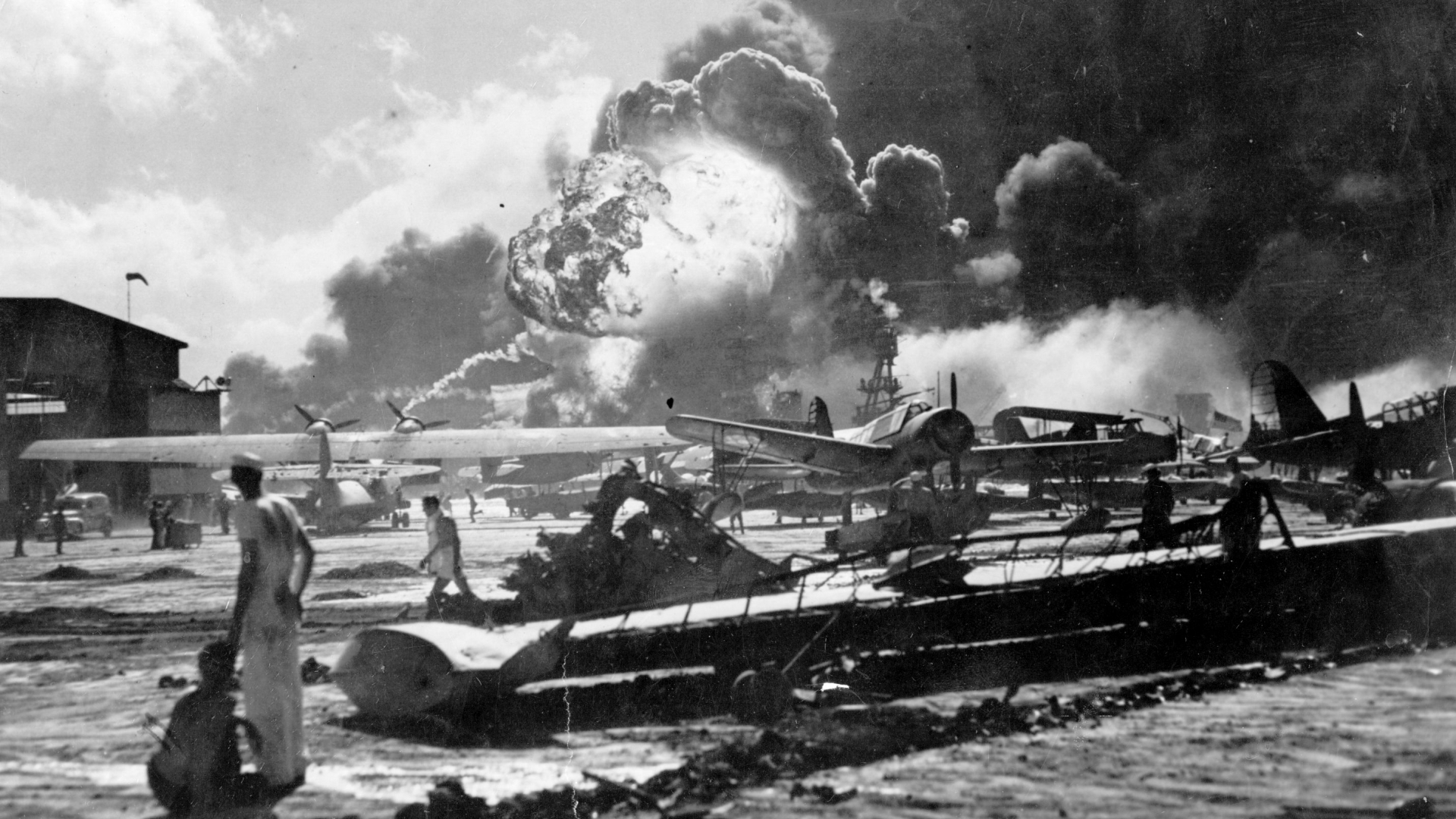 An explosion at the Naval Air Station during the Japanese attack at Pearl Harbor. Sailors stand amid wrecked watching as the USS Shaw explodes in the center background. The USS Nevada is also visible in the middle background, with her bow headed toward the left. (Credit: Fox Photos/Getty Images)
